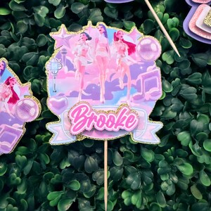May include: A decorative cake topper featuring a group of stylized figures in white outfits, set against a pastel sky background. The topper includes the name "Brooke" in pink script, accented with gold glitter.