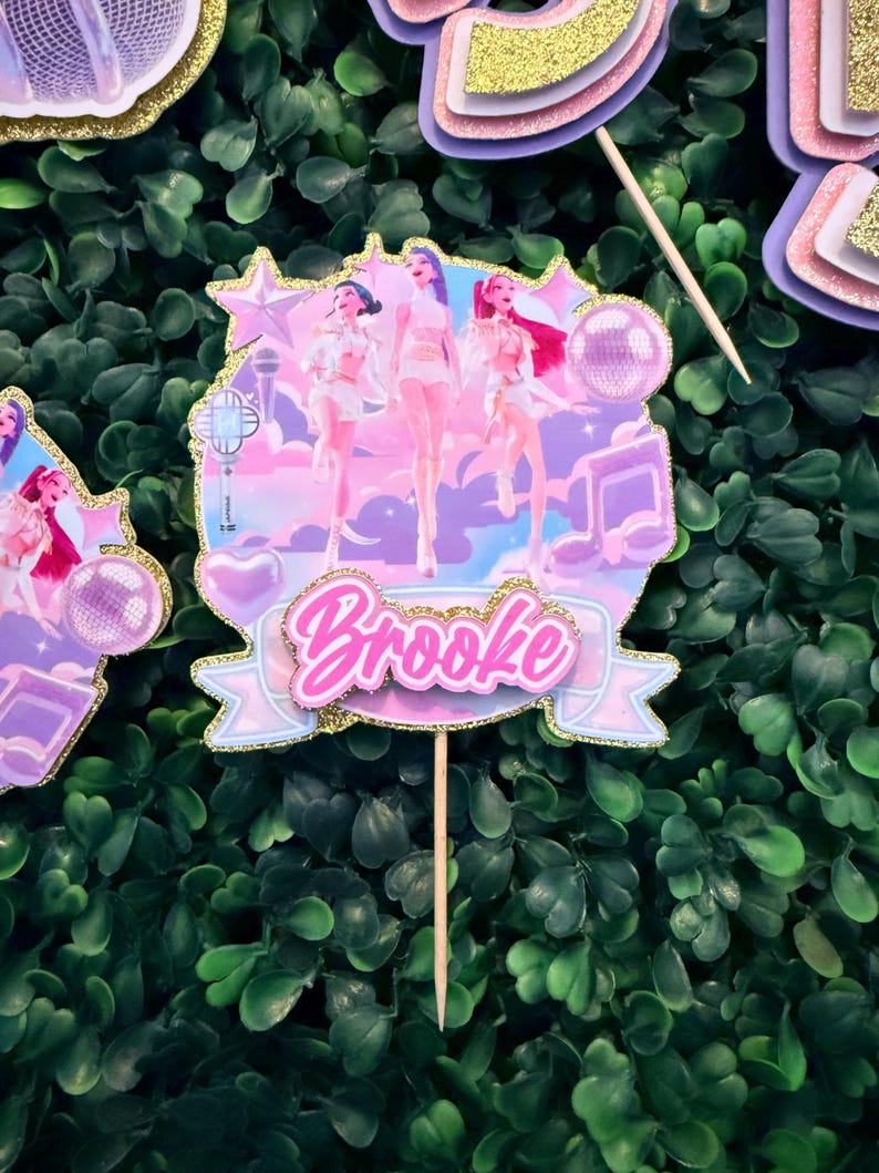 May include: A decorative cake topper featuring a group of stylized figures in white outfits, set against a pastel sky background. The topper includes the name "Brooke" in pink script, accented with gold glitter.
