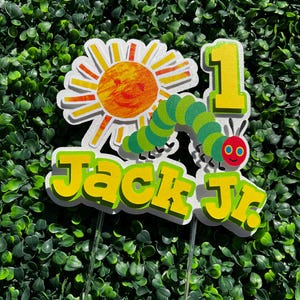 May include: A white, yellow, and green cake topper featuring a smiling sun, a green caterpillar, and the number 1. The text "Jack Jr." is written in yellow letters.