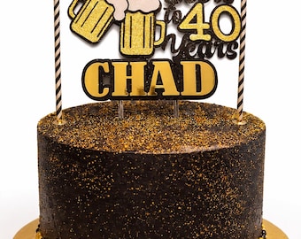 May include: A round, black cake with gold sprinkles and a gold base. The cake is decorated with a topper that says "Cheers & Beers to 40 Years CHAD" and two striped candles.