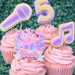 May include: Pink frosted cupcakes with decorative toppers. Toppers include a microphone, a musical note, the number 5, and a banner with the name "Brooke". The toppers have glitter accents and a pop star theme.