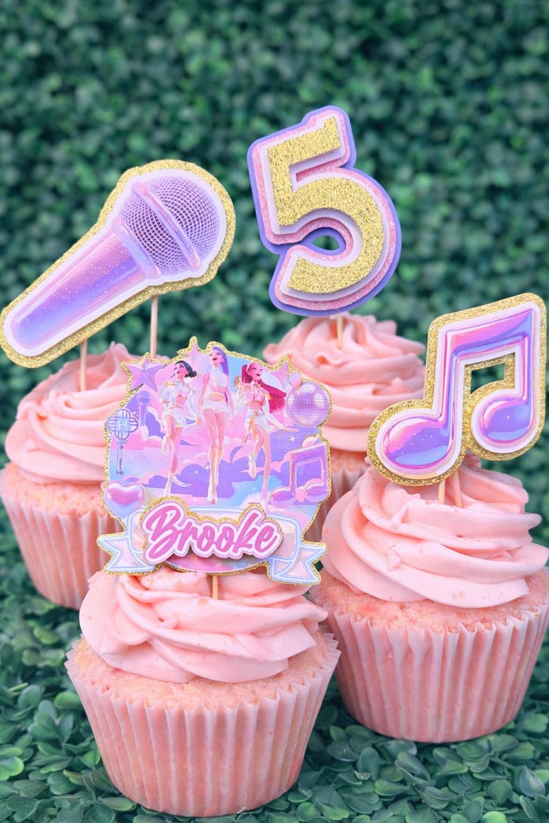 May include: Pink frosted cupcakes with decorative toppers. Toppers include a microphone, a musical note, the number 5, and a banner with the name "Brooke". The toppers have glitter accents and a pop star theme.