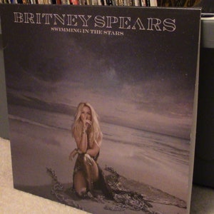Puede incluir: Una portada de álbum de vinilo con Britney Spears. La portada es de color gris oscuro con una foto de Britney Spears sentada en una playa con un cielo estrellado de fondo. El título del álbum "Swimming in the Stars" está impreso en blanco en la portada.