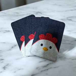 Peut inclure: Trois dessous de verre en tissu denim avec un motif de visage de poulet blanc. Le poulet a une crête rouge et un bec jaune. Les dessous de verre sont disposés en éventail sur une surface en marbre.