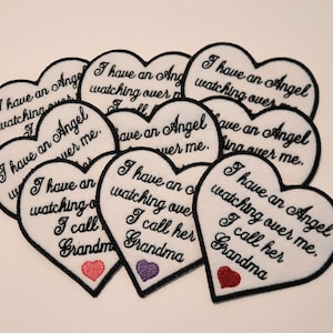 May include: A collection of white felt heart-shaped patches with black embroidery. Each patch features the text "I have an Angel watching over me. I call her Grandma".