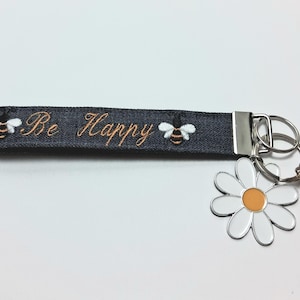 May include: A dark grey keychain wristlet with the embroidered words "Be Happy" and two embroidered bees. A large white daisy charm with an orange centre and a small bee charm are attached to the key rings.