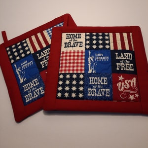 May include: Two red quilted pot holders with a patriotic theme. One pot holder features a patchwork design with the words "Home of the Brave", "Lady Liberty", "Land of the Free", and "USA". The other pot holder features a similar design with the words "Home of the Brave" and "Land of the Free".
