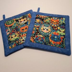 May include: Two blue and white quilted pot holders with a colourful cat print. The cats have bright colours and are surrounded by flowers and paisley patterns.