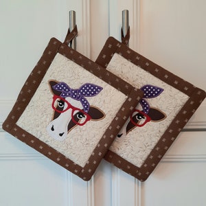 May include: Two brown pot holders with a white background and a cow design. The cow has purple and white polka dot bandana and red glasses.