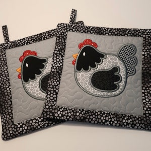 Peut inclure: Deux maniques matelassées grises et noires avec une bordure à pois noirs et blancs. Chaque maniques présente un poulet brodé noir et blanc avec des accents rouges.