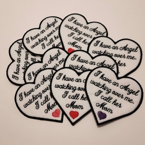 Può includere: Un mucchio di toppe a forma di cuore in feltro bianco con ricami neri. Il testo sulle toppe recita "Ho un angelo che veglia su di me. La chiamo mamma".