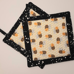 May include: Two black and white pot holders with a bee and honeycomb pattern. The pot holders have a white background with yellow honeycomb and black outlines. The bees are yellow with black stripes. The pot holders have a black border with white splatter paint.