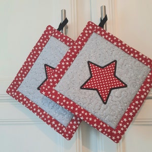 May include: Two red and white quilted pot holders with a large red star with white polka dots in the centre. The pot holders have a black border and a white background with a subtle pattern.