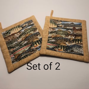 May include: Set of two brown pot holders with a pattern of colourful fish. The pot holders have a tan border and a loop for hanging. "Set of 2" is written below the pot holders.