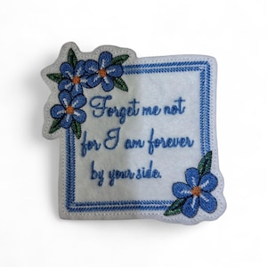 Może przedstawiać: Biała, haftowana naszywka z niebieską obwódką i akcentami kwiatów niezapominajki. Naszywka zawiera haftowany tekst "Forget me not for I am forever by your side" w niebieskim kolorze.