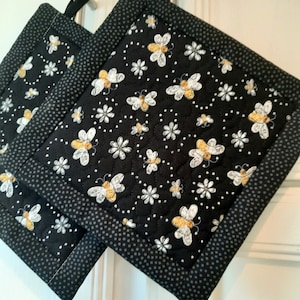 Peut inclure: Deux maniques de cuisine matelassées noires avec une bordure à pois blancs. Les maniques présentent un fond noir avec un motif répétitif de marguerites blanches et d'abeilles jaunes et blanches.