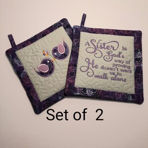 Peut inclure: Ensemble de deux maniques carrées matelassées. L'une présente des oiseaux violets brodés avec une fleur, l'autre le texte brodé "A Sister is God's way of proving He doesn't want us to walk alone." Les maniques ont une bordure à motifs violets et blancs.