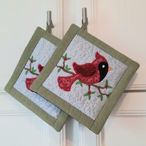 May include: Two quilted pot holders with a green border and a white background. Each pot holder features an embroidered cardinal bird with red and black accents, perched on a branch with green leaves and red berries.