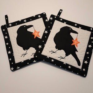 May include: Two black and white quilted pot holders with a black silhouette of a crow holding a star in its beak. The pot holders have a black border with white stars.