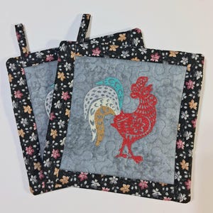 Peut inclure: Deux maniques carrées matelassées avec un motif de coq. Le coq est rouge avec une queue turquoise, blanche et dorée. Les maniques ont un fond gris et sont bordées d'un tissu floral noir. Chaque manique a une boucle pour suspendre.