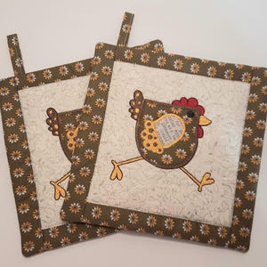 May include: Two brown and yellow fabric pot holders with a white background and a brown chicken design. The chicken has a red comb and a yellow body with brown accents. The text on the chicken reads "For after all, the best things in life are free. The pleasure to give and the joy to receive."