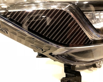 Pode incluir: Close-up de um conjunto de faróis de carro com um detalhe em fibra de carbono. A trama de fibra de carbono preta e cinza é visível dentro de uma carcaça clara e geométrica. O farol tem uma lente transparente e um suporte de montagem preto.