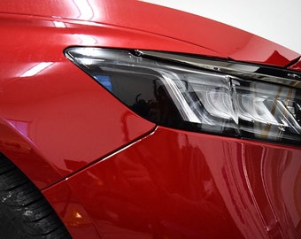 May include: A bright red car exterior featuring a sleek, angular headlight assembly with integrated LED lighting elements. The glossy paint finish reflects light, highlighting the sharp body lines and the transition between the fender and hood.