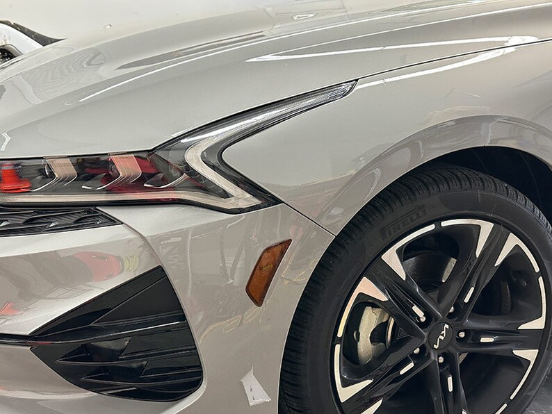 May include: A silver car with black wheels and a black front bumper. The car has a silver paint job and black accents. The headlights are on and the car is parked in a garage.