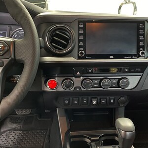 Op de afbeelding: Het interieurdashboard van een voertuig met een zwart stuurwiel met bedieningselementen, een centraal JBL-touchscreen, ronde ventilatieopeningen en een opvallende rode startknop naast de knoppen voor de klimaatregeling.