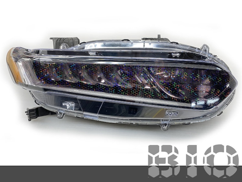May include: A clear plastic car headlight with a black housing and a holographic film covering the lens. The film has a rainbow iridescent effect.