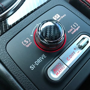 May include: Close-up of a car's dashboard with a black carbon fiber knob and a silver button labeled "SI-DRIVE". The knob has a red ring around it and the button has a red inscription. There are also buttons labeled "AUTO", "MANU", and "C.DIFF + -" with red inscriptions.