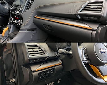 May include: Close-up of a car's interior with black and orange accents. The image shows the dashboard, steering wheel, and air vents. The steering wheel has a Subaru logo.