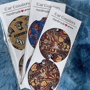 May include: Three sets of handmade car coasters featuring different college logos. The coasters are round and have a variety of colors and designs. The logos include the University of Florida, Florida State University, and the University of Central Florida.