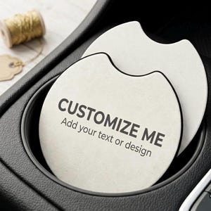 May include: Two off-white car coasters with black trim, one inside a car cup holder. The coasters have the text "CUSTOMIZE ME" and "Add your text or design" printed on them. A spool of gold thread and a blank tag are in the background.