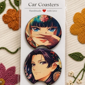 以下が含まれることがあります： Car Coasters Handmade with loveと書かれた白い台紙に透明なプラスチック袋で包装された、2枚の円形カーコースター。上のコースターには青い目と黒髪のアニメ風キャラクターが描かれ、下のコースターには紫色の目と黒髪のアニメ風キャラクターが描かれています。