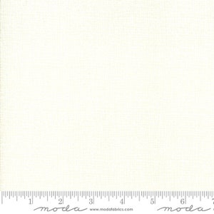 May include: Cream-colored fabric with a subtle woven texture. The fabric is a solid, neutral color, and a ruler is visible along the bottom edge. The fabric is from Moda Fabrics.