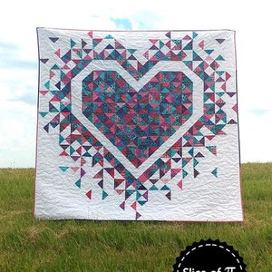May include: A quilt with a large pink and blue heart design. The heart is surrounded by smaller triangles in shades of pink, blue, and purple. The quilt is displayed on a grassy field with a blue sky in the background. The text "Exploding Heart" is at the top of the image and "Slice of Pi Quilts" is at the bottom.