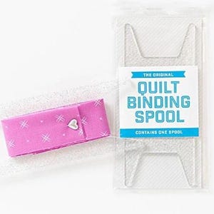May include: A pink fabric spool holder with a heart-shaped clasp and white crosshatch pattern. The holder is next to a clear plastic package with the text "The Original Quilt Binding Spool".