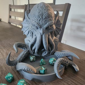 以下が含まれることがあります： クトゥルフ風のクリーチャーの頭部が彫刻された、グレーの3Dプリント製ダイストレイ。触手が円形のベースの周りに伸びており、ターコイズブルーと白のマーブル模様の多面体ダイスがトレイの内外にいくつか散らばっています。