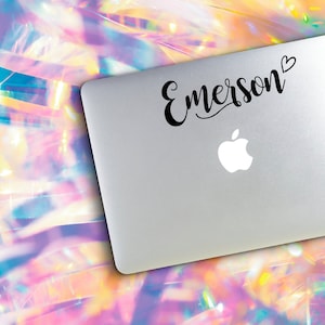 May include: A silver laptop with a black sticker that says "Emerson" with a heart next to it. The laptop is on an iridescent background.