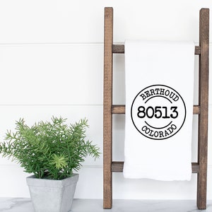 May include: White kitchen towel with a black circle design that says "Berthoud Colorado 80513" hanging on a wooden ladder.