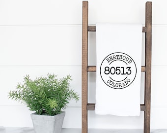 Peut inclure: Une serviette de cuisine blanche avec un motif circulaire noir qui dit "Berthoud Colorado 80513" accrochée à une échelle en bois.