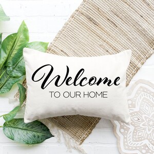 Puede incluir: Una almohada blanca con texto negro que dice "Bienvenido a nuestro hogar". La almohada está sobre una alfombra tejida con un fondo blanco.