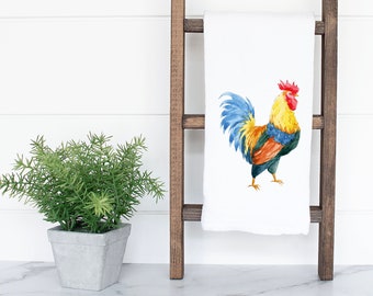 May include: A white kitchen towel with a watercolor illustration of a colorful rooster. The rooster has a red comb, yellow body, blue tail feathers, and orange and green accents.