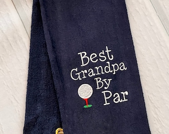 May include: A navy blue golf towel featuring white embroidered text that reads Best Grandpa By Par, accompanied by a small embroidered graphic of a white golf ball on a red tee with a patch of green grass and a gold metal grommet with a hook.