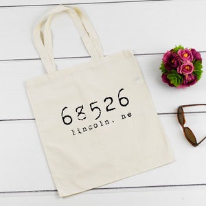 Op de afbeelding: Een witte canvas tote bag met zwarte tekst die "68526 lincoln, ne" zegt.