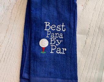 May include: A royal blue hand towel featuring white embroidered text that reads Best Papa By Par, accompanied by a small white golf ball on a red tee with a green base. The towel includes a gold-toned metal hook attached to the bottom edge.