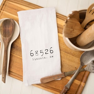 May include: White kitchen towel with the text "68526 lincoln, ne" printed on it. The towel is lying on a wooden cutting board with kitchen utensils.