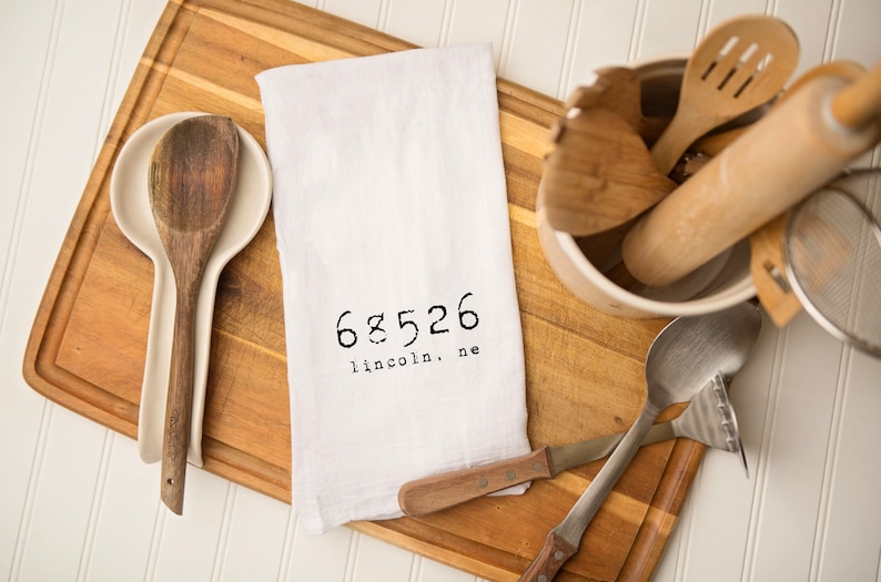 May include: White kitchen towel with the text "68526 lincoln, ne" printed in black. Wooden spoons, a rolling pin, and other kitchen utensils are arranged on a wooden chopping board.