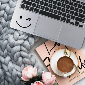 Puede incluir: Un portátil plateado con una pegatina de carita sonriente negra, una manta de punto grueso gris, un libro rosa, una taza de café y flores rosas. La taza de café es blanca con un borde dorado y una cuchara dorada.